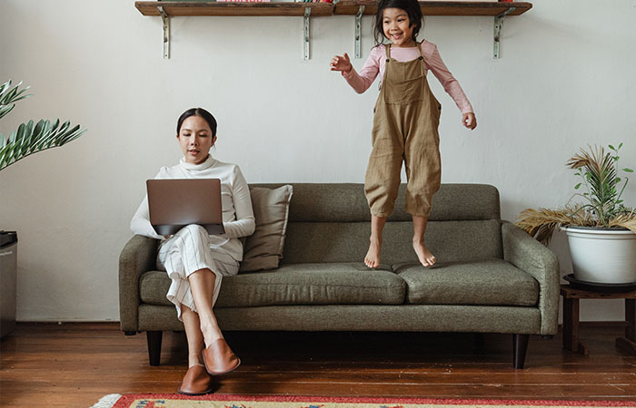 Image of woman trying to work with daughter jumping on the sofa next to her