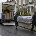A white Luton removals van with its tail lift down on a South London terraced street as two movers carry a wrapped sofa.