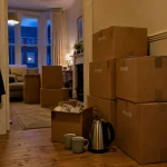 Labelled moving boxes stacked in a hallway at dusk, kettle and mugs kept out, the night before a move done properly