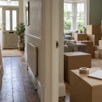 Stacks of cardboard moving boxes in the hallway and front room of a terraced house, with tape and a notepad to count them.